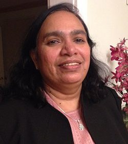 A smiling woman with dark hair and a necklace, standing indoors near a pink flower decoration.